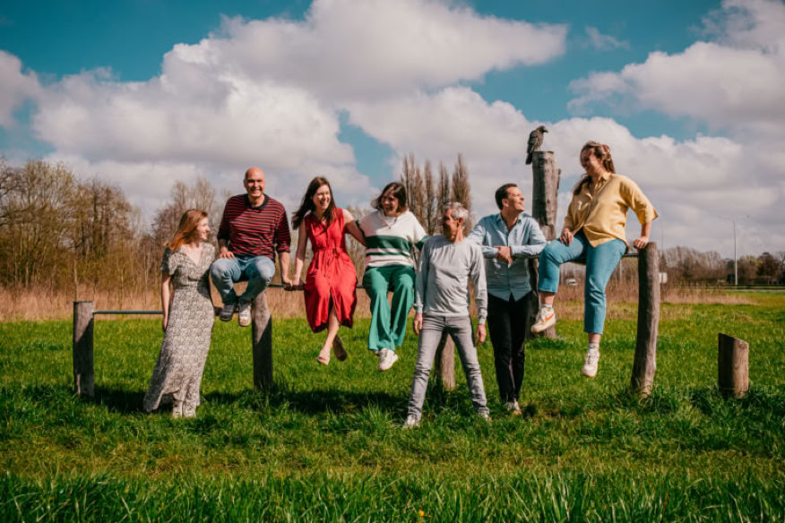 Een groep van zeven mensen zit en staat op een houten hek in een grasveld onder een blauwe lucht met wolken.