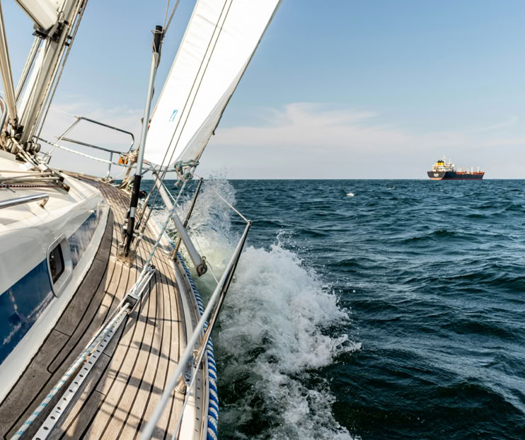 Zeilboot op zee met golven en een groot schip in de verte onder een blauwe hemel.
